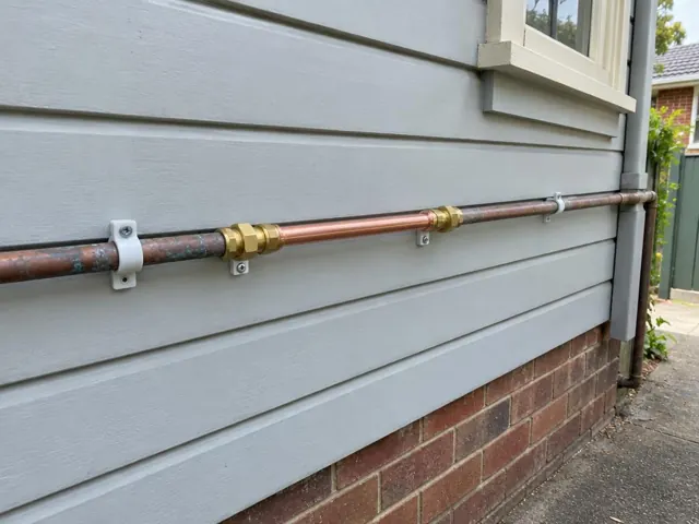 Repaired copper water pipe running along exterior weatherboard wall above brickwork in Arncliffe